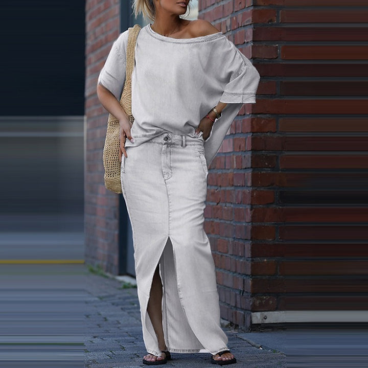 Woman in a light gray off-shoulder top and white pants standing against a brick wall.