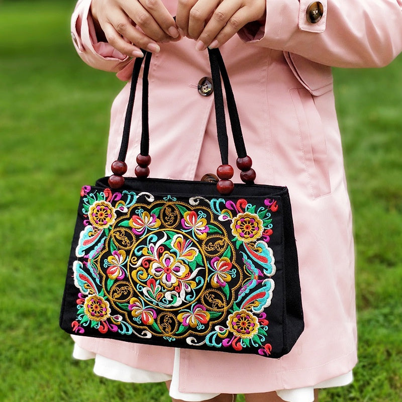 Person holding a black handbag with colorful floral embroidery against a green grass background