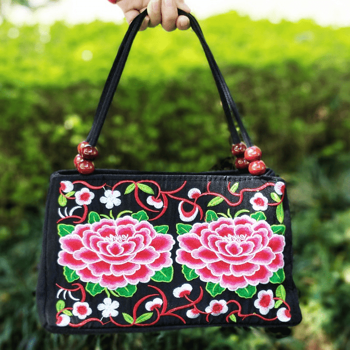 Black handbag with pink floral embroidery held by a hand against a green blurred background