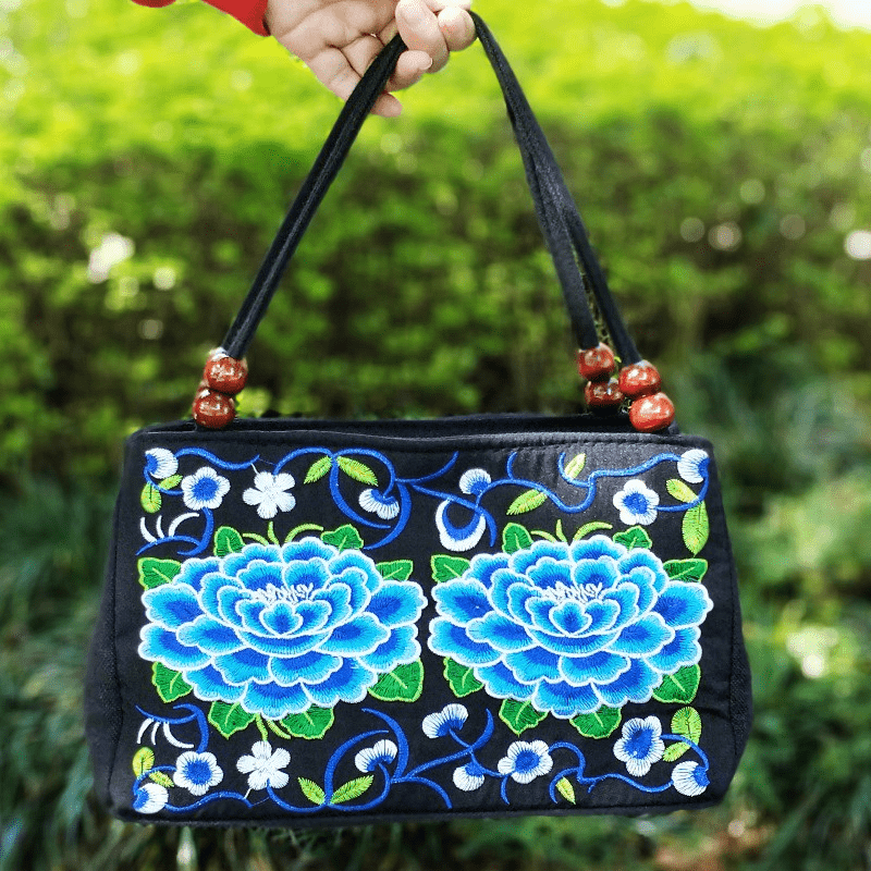 Black handbag with blue floral embroidery held by a hand against a green outdoor background