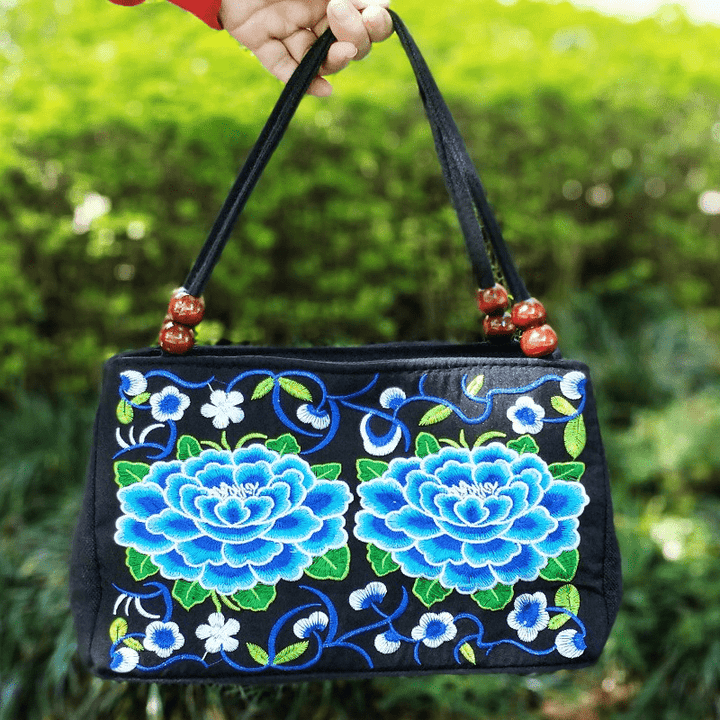 Black handbag with blue floral embroidery held by a hand against a green outdoor background