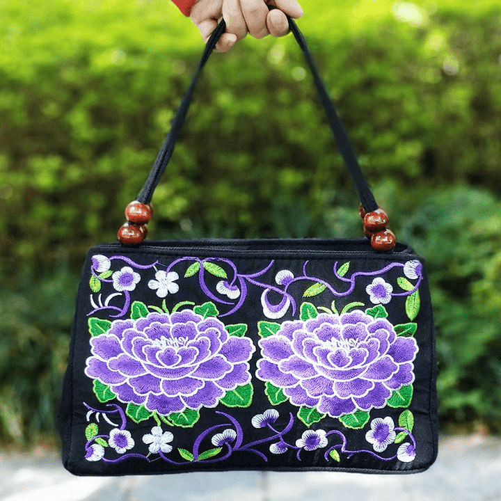 Black handbag with purple floral embroidery held by a hand against a green outdoor background