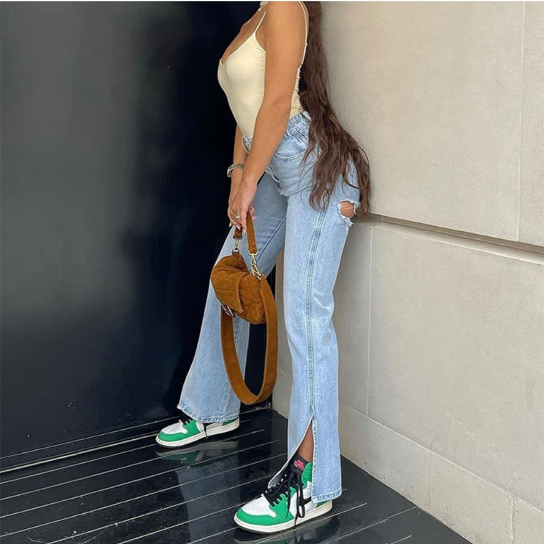 Person wearing a beige top, blue jeans, and green sneakers holding a brown handbag against a black wall.