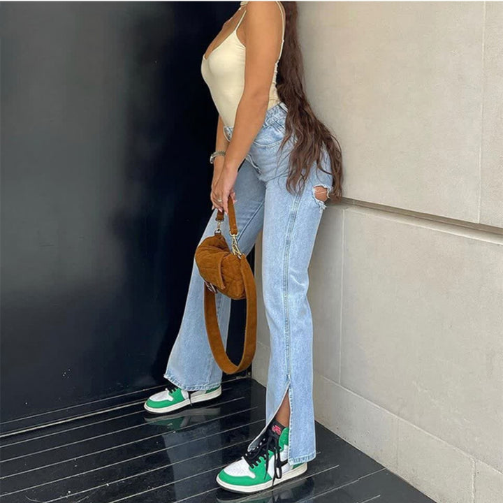 Person wearing a beige top, blue jeans, and green sneakers holding a brown handbag against a black wall.