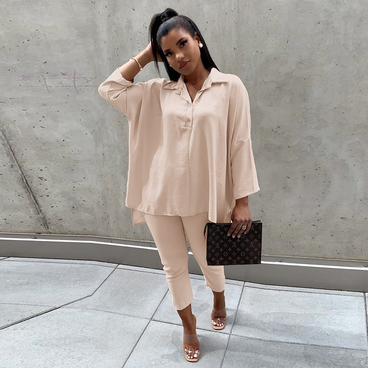 Woman in beige outfit holding a black clutch against a concrete wall.
