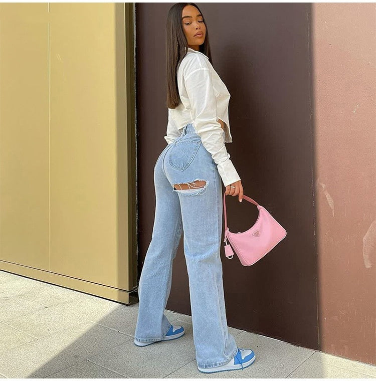 Person wearing light blue jeans and a white top, holding a pink handbag against a brown wall.