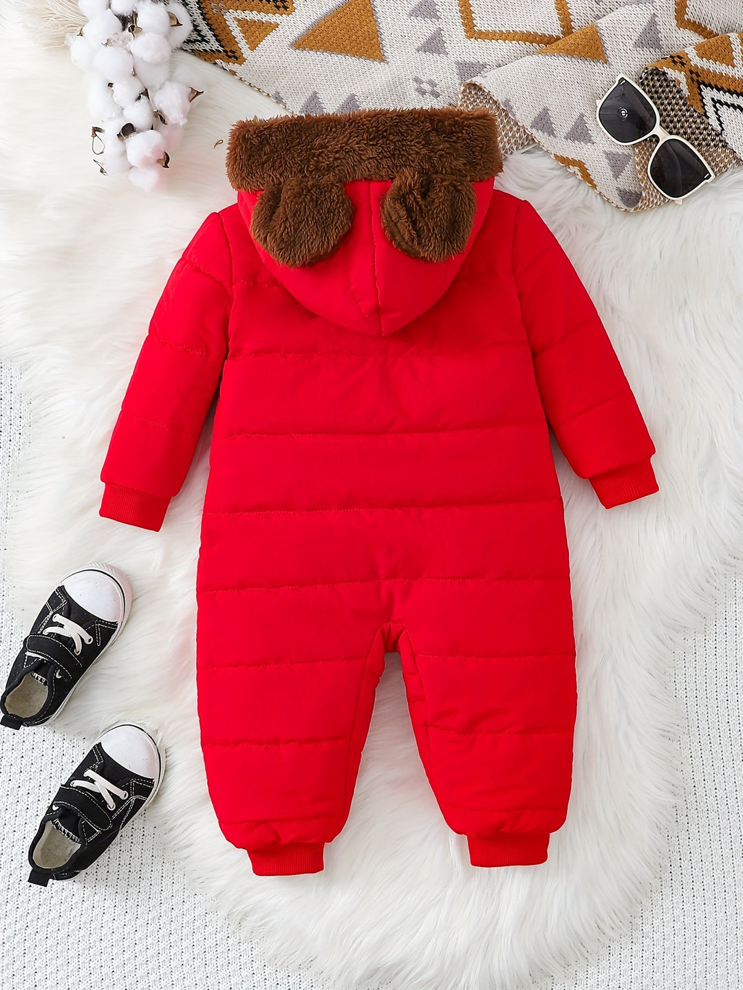 Red baby snowsuit with brown hood on a white surface with black shoes and sunglasses.