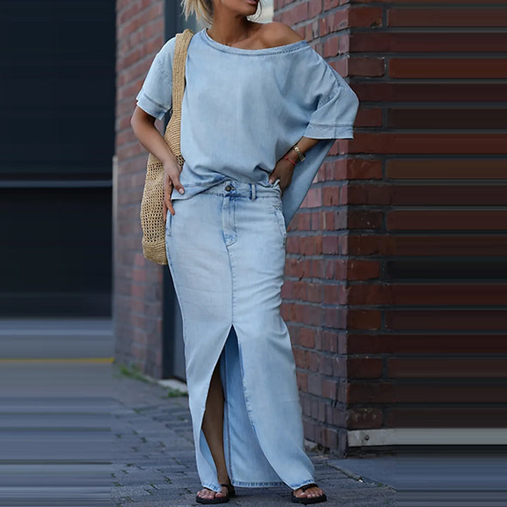 Woman wearing a light blue off-shoulder top and jeans, standing against a brick wall.
