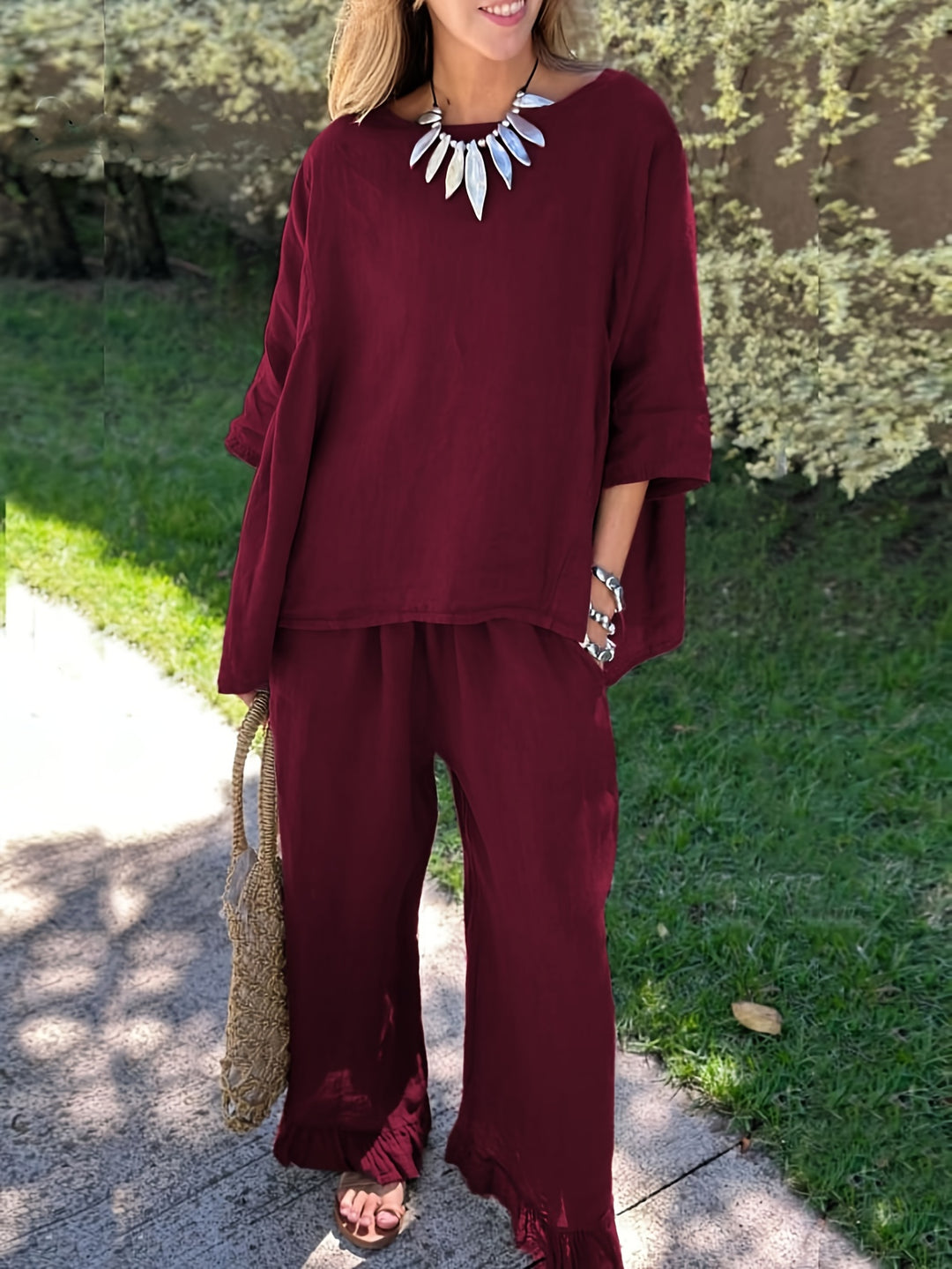 Woman wearing a burgundy outfit with ruffled pants and holding a straw bag outdoors.