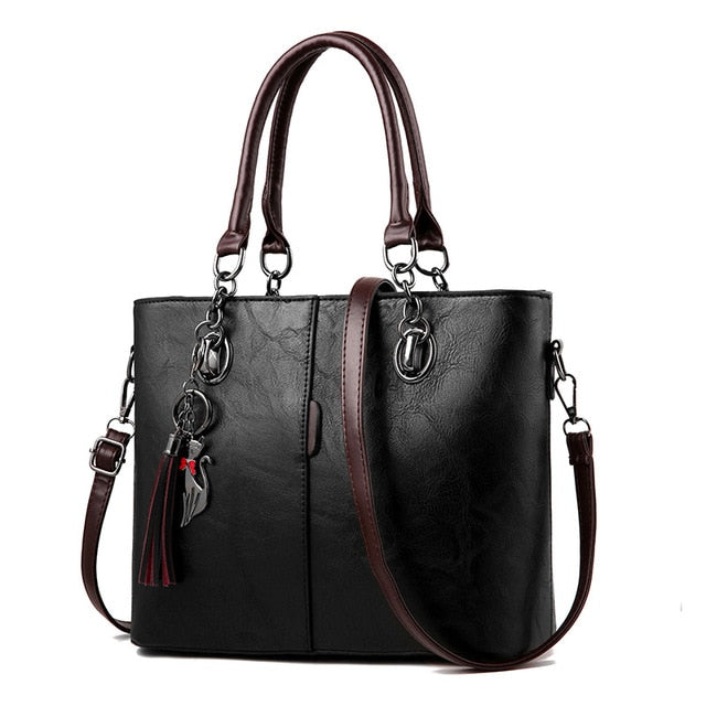 Black leather tote bag with structured front, top handles, crossbody strap, tassel charm