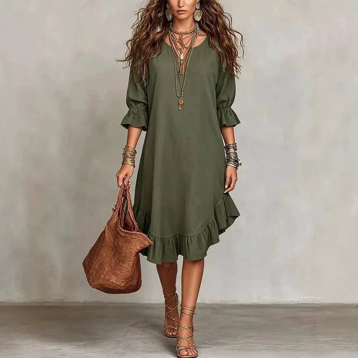 Woman wearing an olive green dress holding a brown bag against a plain background