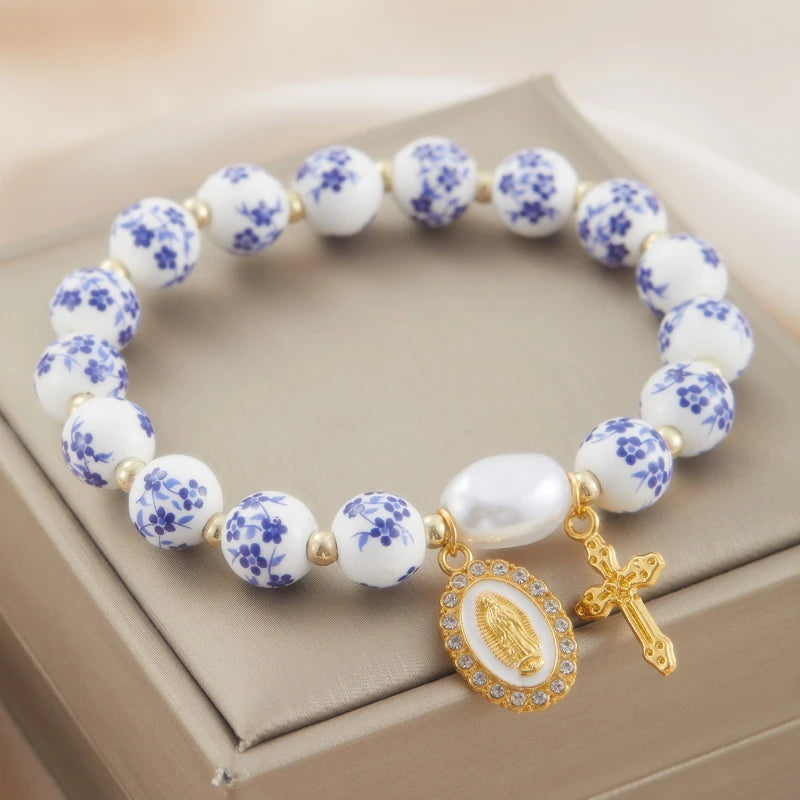 Bracelet with white beads and blue floral patterns, featuring a gold charm and cross on a beige surface.