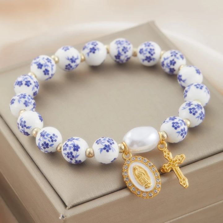 Bracelet with white beads and blue floral patterns, featuring a gold charm and cross on a beige surface.