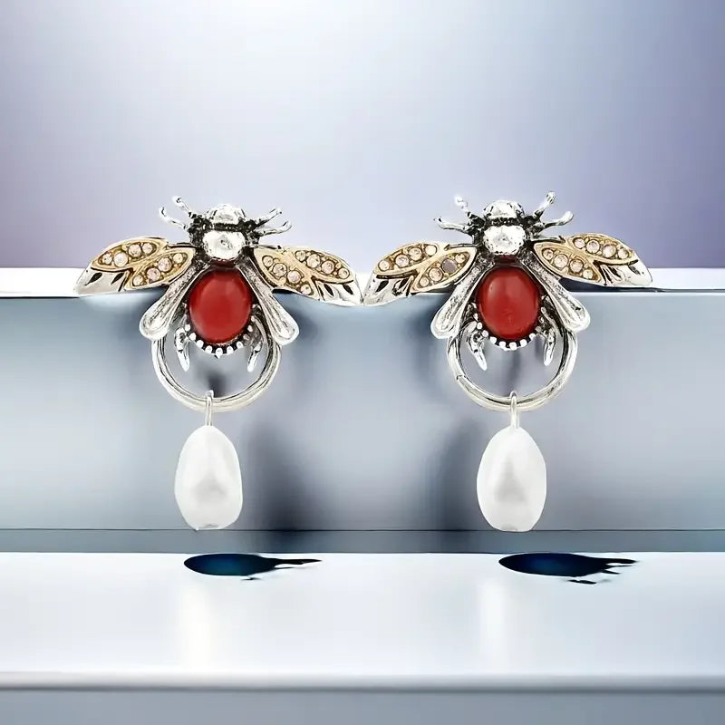 Bee-shaped earrings with red gemstones and pearl drops on a reflective surface.