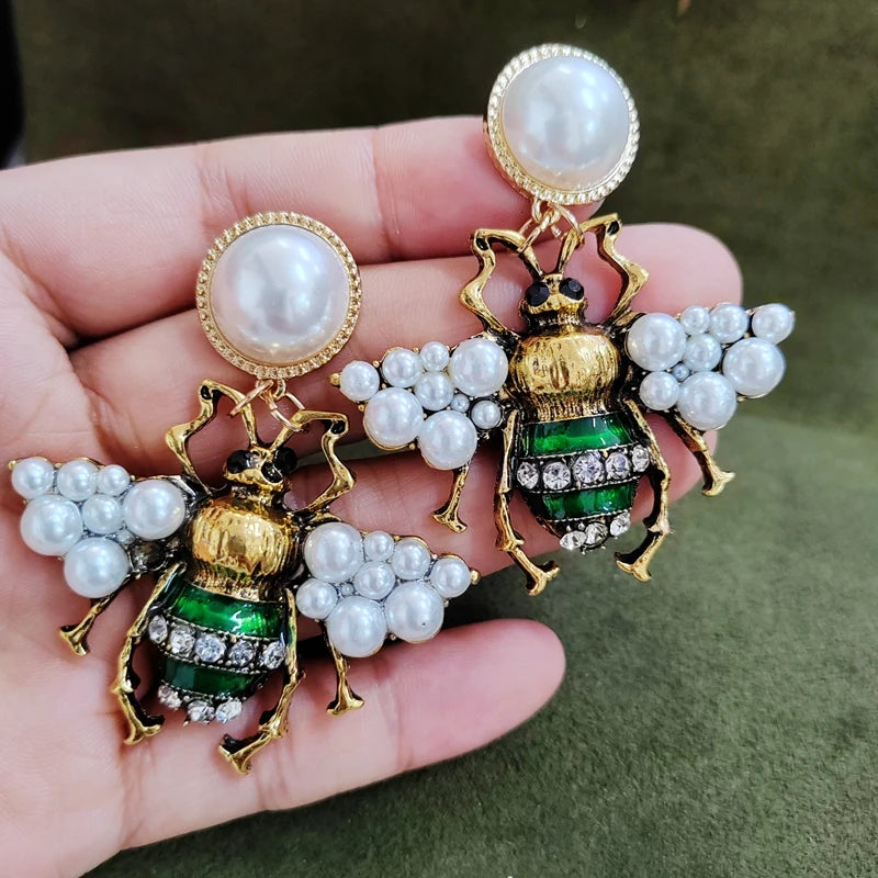 Gold and green insect-shaped earrings with pearl accents held in a hand against a green background.