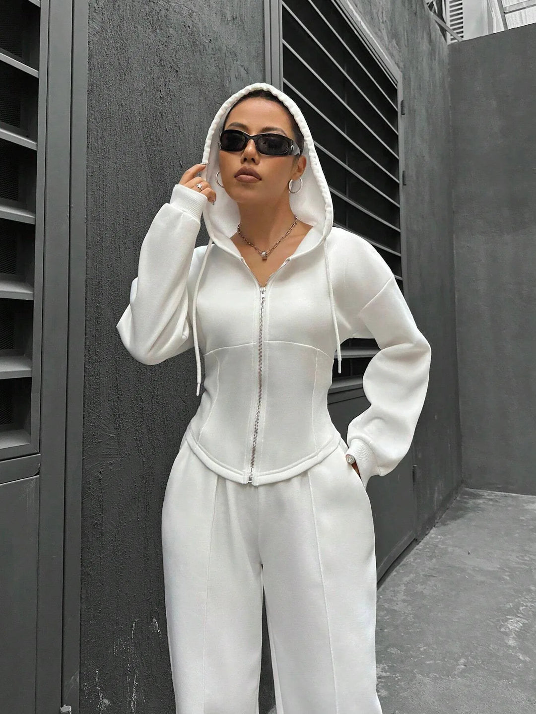 Person wearing a white tracksuit and sunglasses standing against a gray wall.