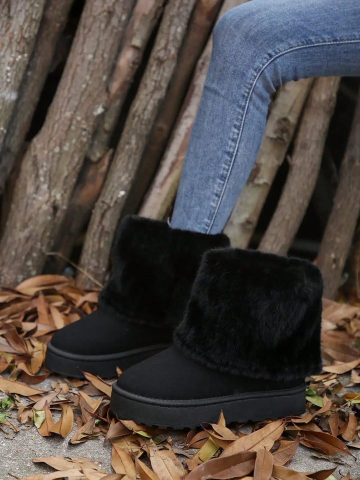 Black faux-fur cuffed ankle boots worn with blue jeans, set outdoors on a ground covered in dry leaves in front of a rustic wood log wall.
