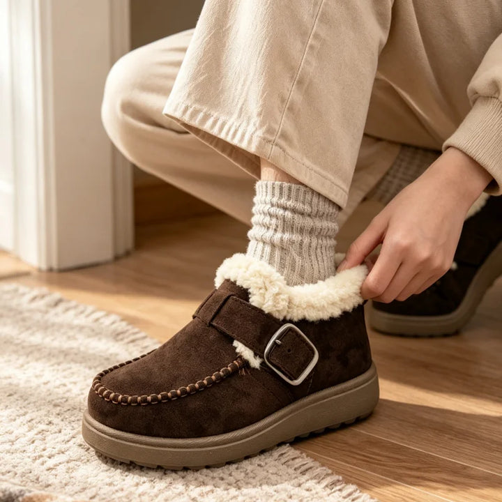 A person wearing beige pants and a cream ribbed sock is shown from the knees down, putting on a dark brown suede slipper with a fluffy cream sherpa lining and a silver buckle strap. The slipper is placed on a light-colored rug over a wooden floor.