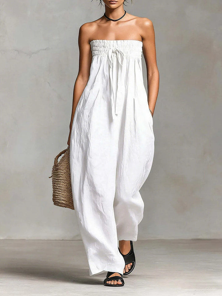 Woman wearing a strapless white dress holding a woven bag against a plain background