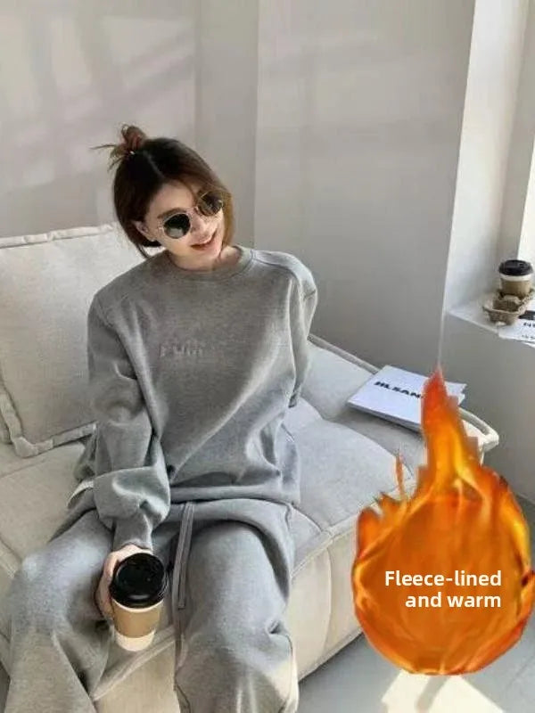 A woman wearing a light gray fleece lounge set sits on a beige sofa, holding a takeaway coffee cup. The set includes a crewneck sweatshirt and matching pants.
