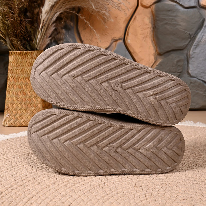 A pair of taupe-brown slip-on slippers shown from the sole, stacked on a woven beige rug with a basket and dried grass in the background.