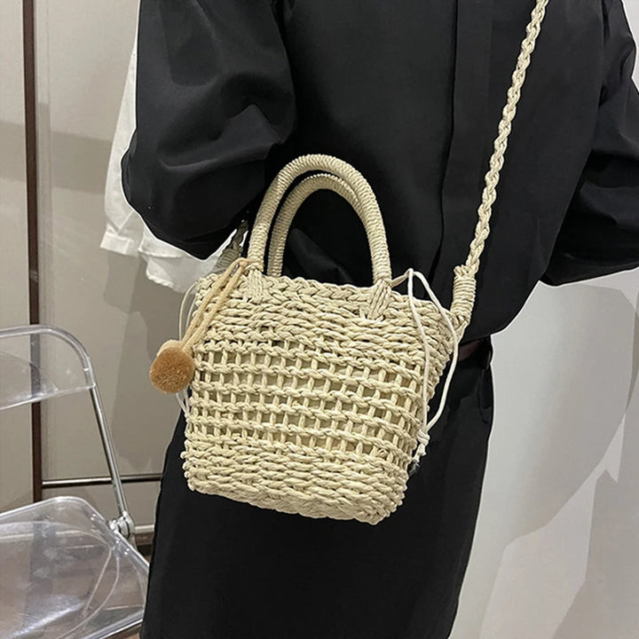 Beige knit straw bucket crossbody with pom-pom charm, tie closure, and long strap