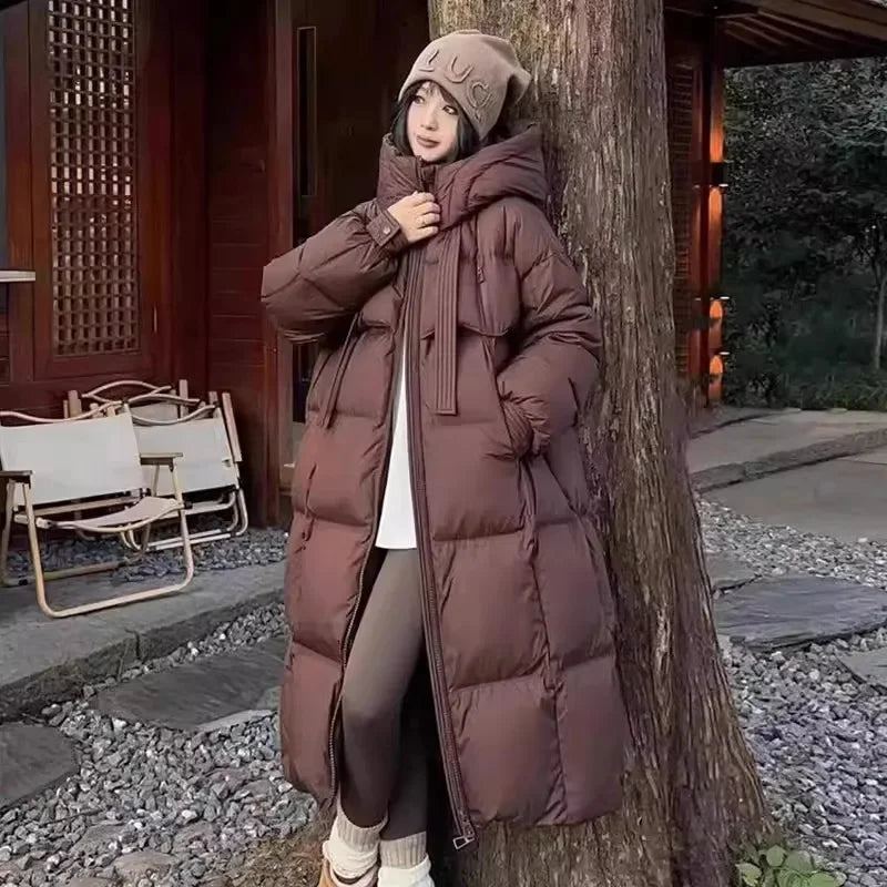 Person wearing a long brown puffer coat and beige hat leaning against a tree outdoors.