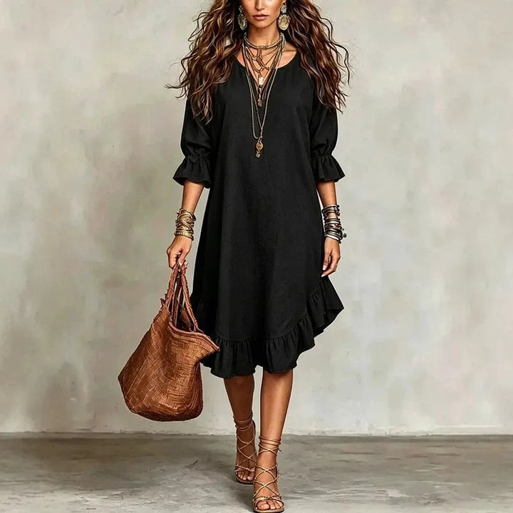 Woman wearing a black dress with ruffled sleeves and holding a brown leather bag against a plain background.