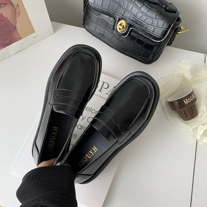 Black loafers with 'HACU' branding on a white surface with a black handbag and cup in the background.