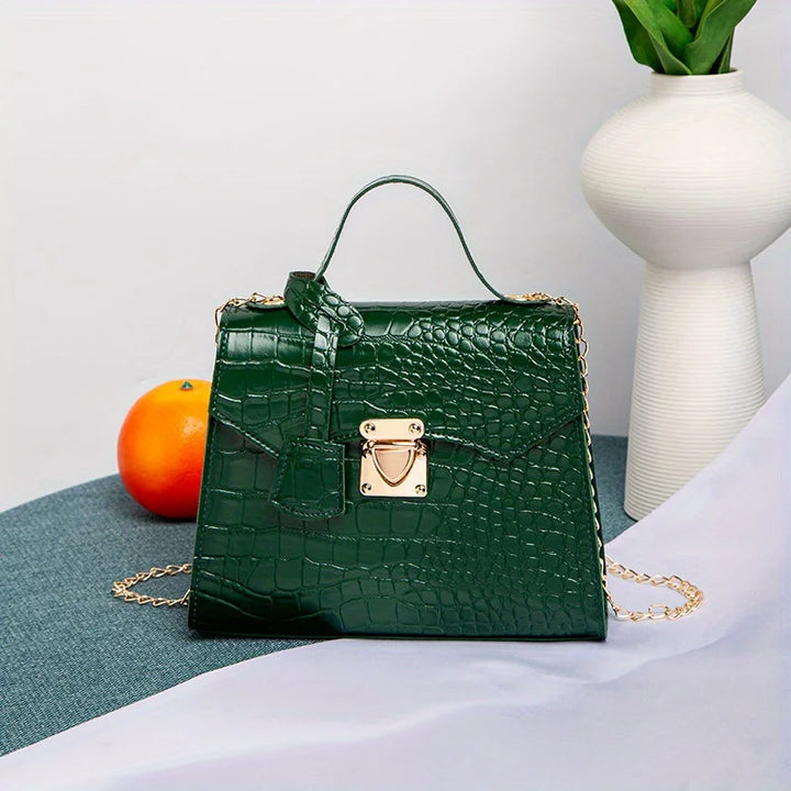 Green crocodile-patterned handbag with gold chain on a white surface with a vase and fruit in the background.
