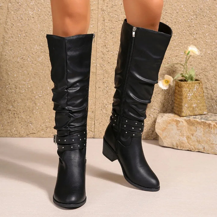 A pair of black faux-leather slouchy knee-high boots with studded straps and low block heels, shown on a model's legs against a textured beige wall and stone decor.