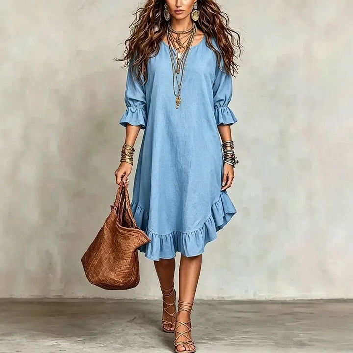 Woman wearing a blue dress holding a brown bag against a plain background