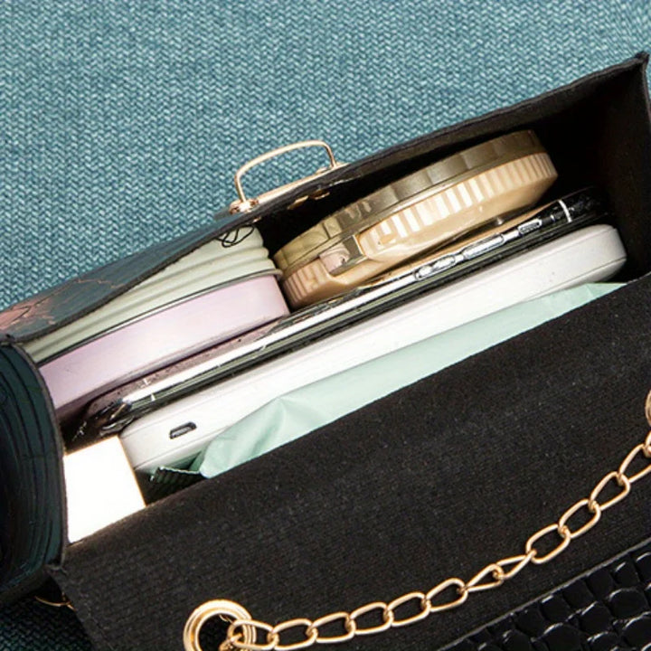 Black handbag with gold chain strap containing makeup items on a textured blue surface