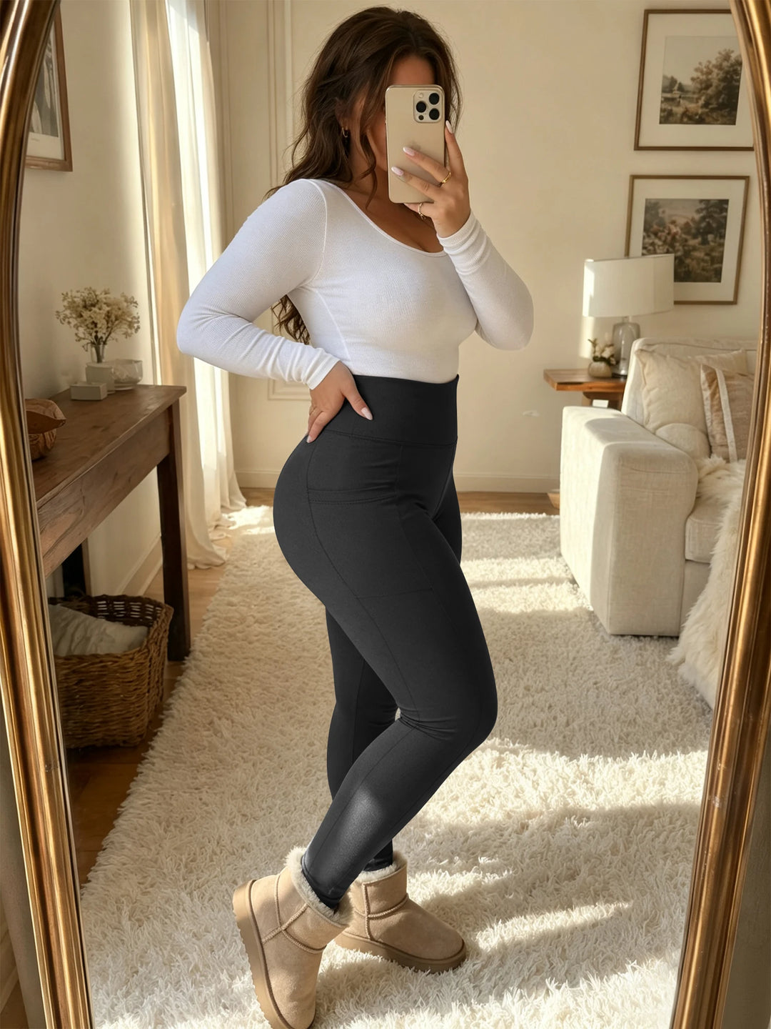 A woman takes a mirror selfie in a cozy living room, wearing a fitted white long-sleeve top, black leggings, and beige ankle boots with faux-fur trim.