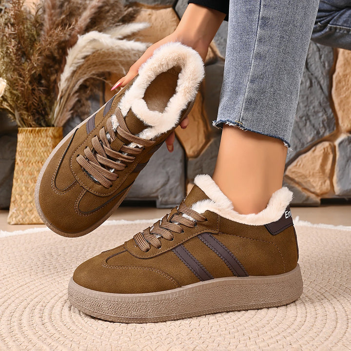 A person wearing brown suede winter boots with white faux-fur lining and dark brown side stripes, standing on a woven rug with dried plants in the background.