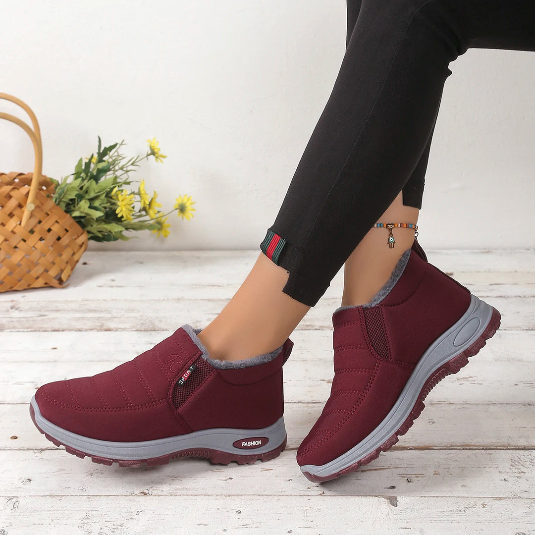Maroon slip-on shoes with gray soles worn by a person on a light wooden floor.