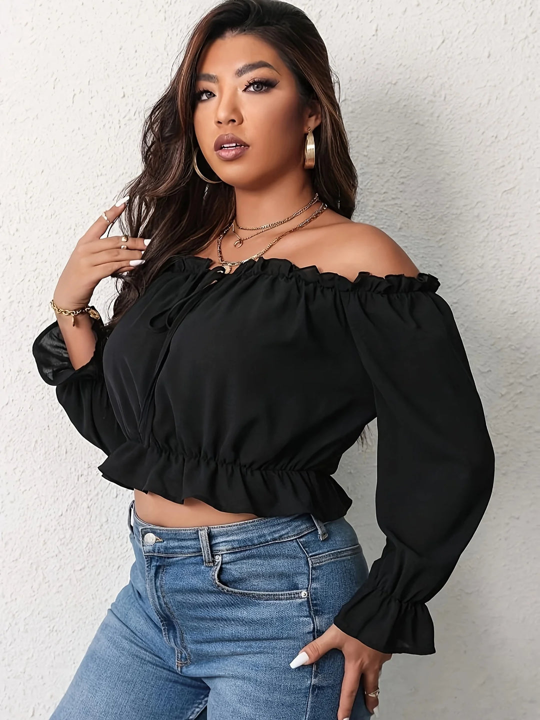 Woman wearing a black off-shoulder top and blue jeans against a white wall.
