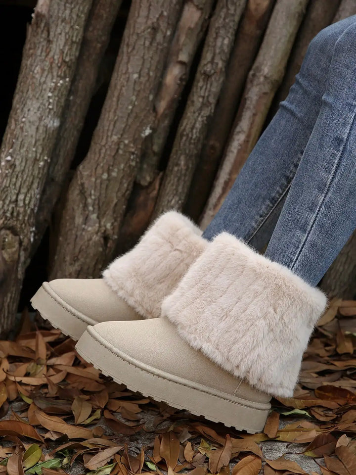 Beige suede ankle boots with fluffy cream faux fur trim, shown on a person wearing blue jeans standing on a bed of fallen leaves in front of a wood wall.