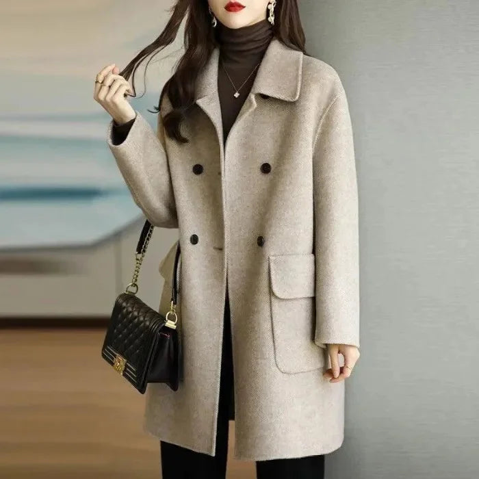 Woman wearing a beige coat with black buttons, holding a black handbag against a neutral background.