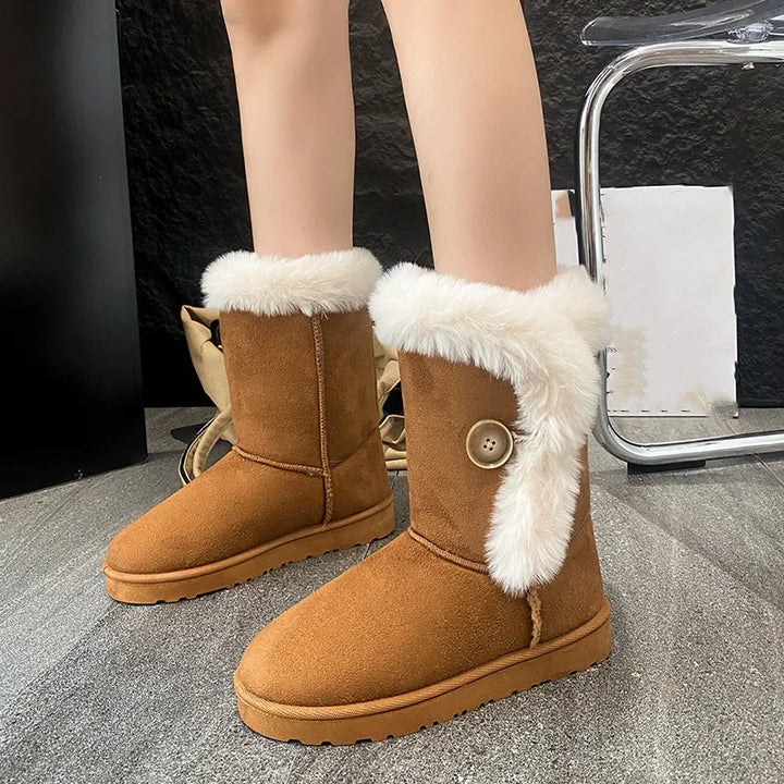 A person wearing tan suede-like ankle winter boots with thick white faux-fur trim and a decorative button detail on the side, standing on a gray floor.