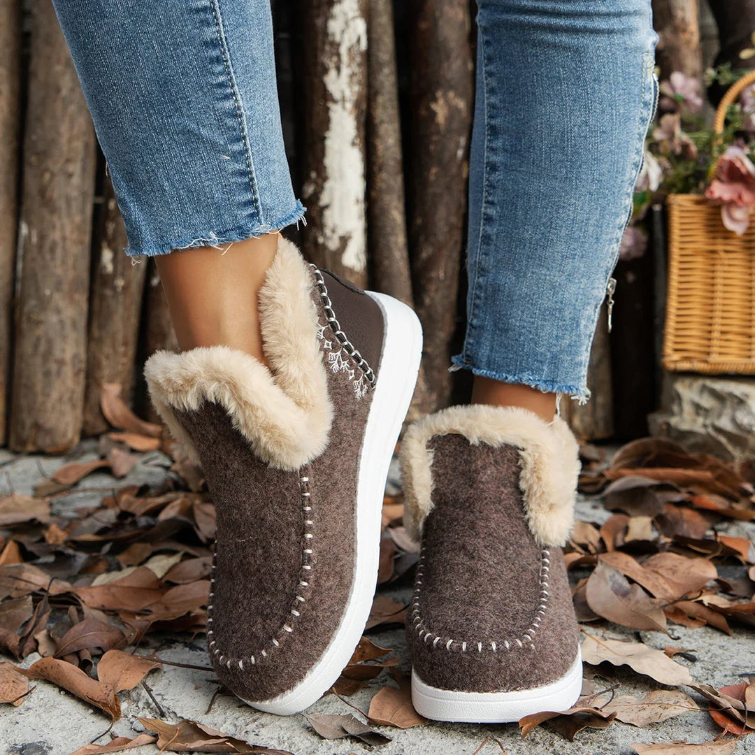 Brown slip-on boots with fur trim worn with blue jeans on a leaf-covered ground.