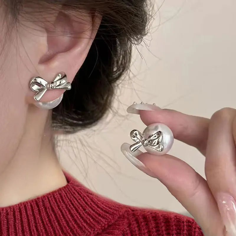 Hand wearing a decorative earring with a bow design on a neutral background