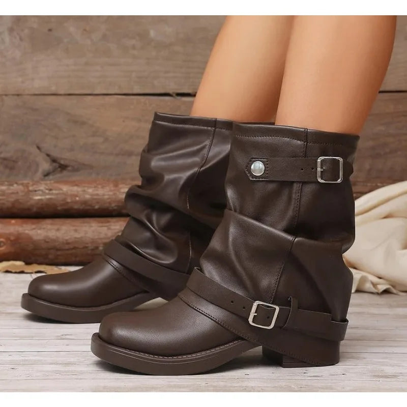 A pair of dark brown faux-leather slouchy ankle boots with buckle straps, shown on a model's feet against a rustic wood background.