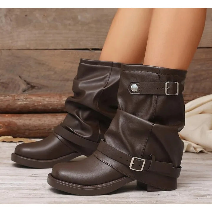 A pair of dark brown faux-leather slouchy ankle boots with buckle straps, shown on a model's feet against a rustic wood background.