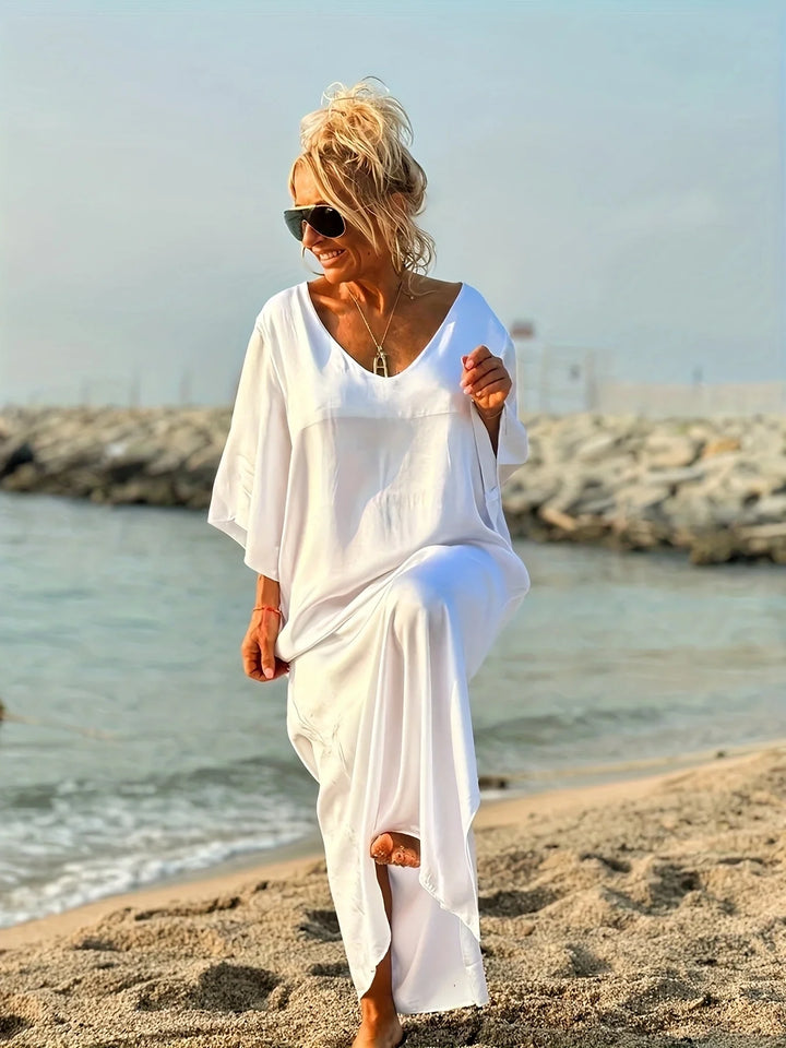 Woman in a white dress walking on a beach with sunglasses and a necklace.