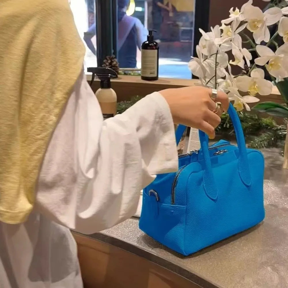A person wearing a white top and yellow head covering holds the handle of a bright blue structured handbag on a countertop in a shop setting, with white flowers and bottles in the background.