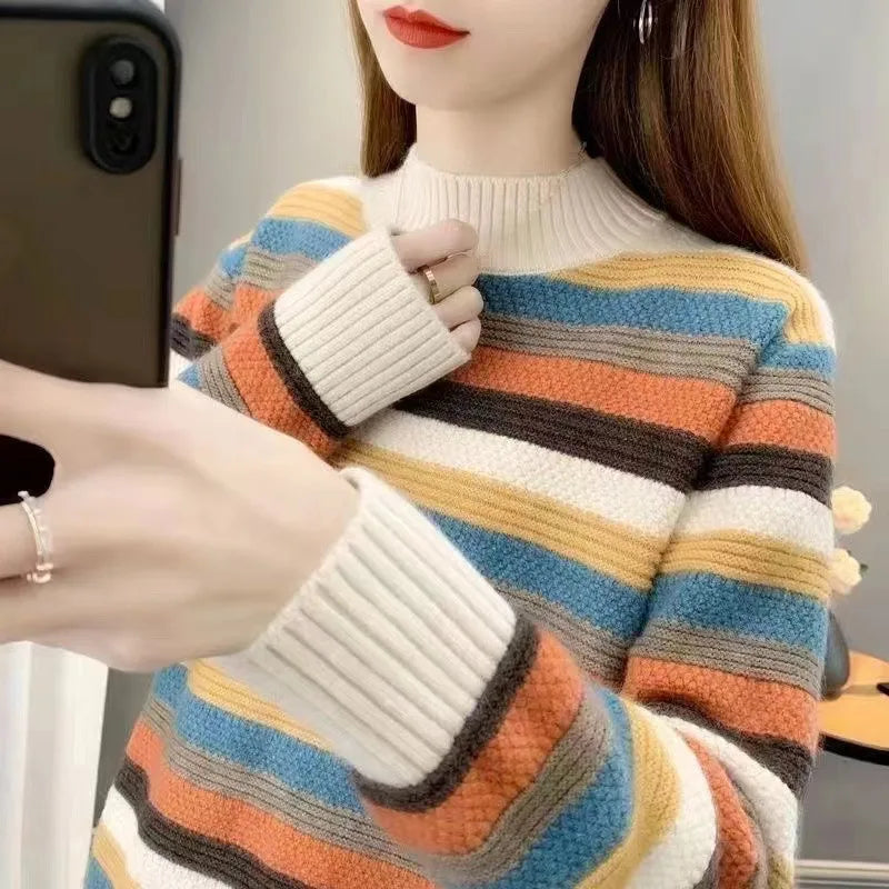 A woman takes a mirror selfie showing off a cozy, multicolored striped knit sweater. The sweater features horizontal stripes of cream, mustard yellow, orange, light blue, and dark brown, with thick cream ribbed cuffs and a matching ribbed mock neck.