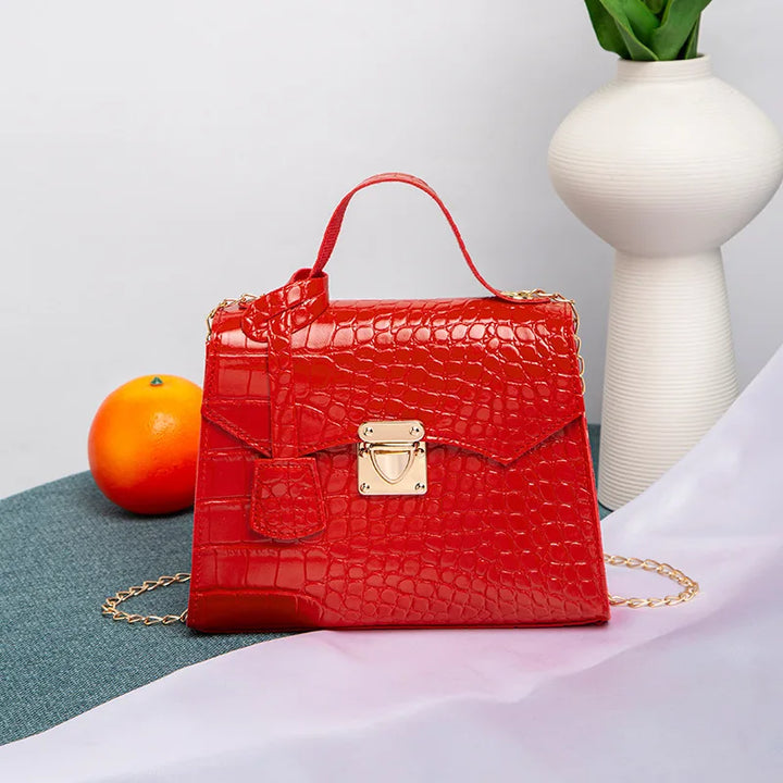 Red handbag with gold accents on a surface with a white vase and orange in the background