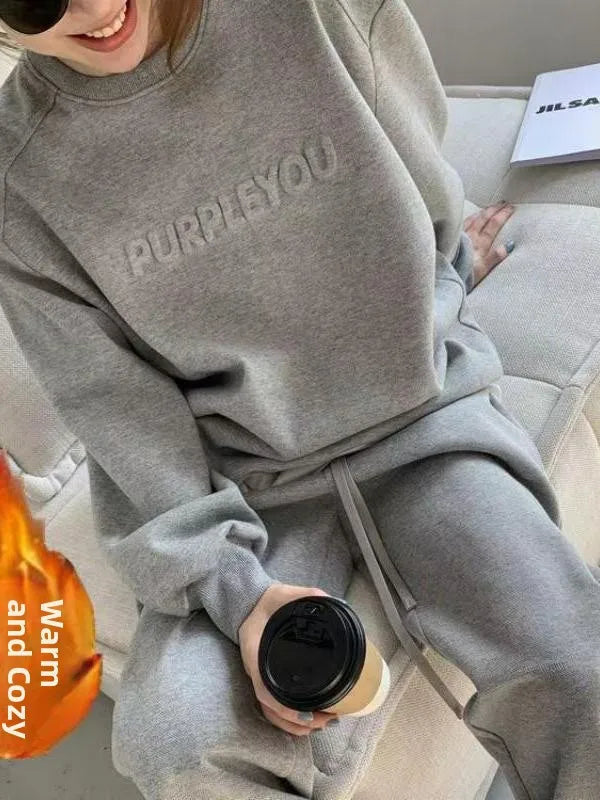 A person wearing a light gray matching sweatshirt and sweatpants set, sitting on a light-colored couch and holding a takeaway coffee cup. The sweatshirt has the word 'PURPLEYOU' embossed on the chest.