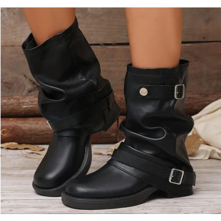 A pair of black faux-leather ankle boots with draped straps and silver buckle details, shown on a model's feet against a rustic wooden background.